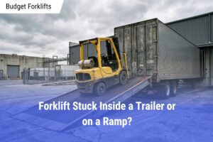 A yellow warehouse forklift stuck partway up a metal loading ramp leading into a closed box trailer at an industrial dock.